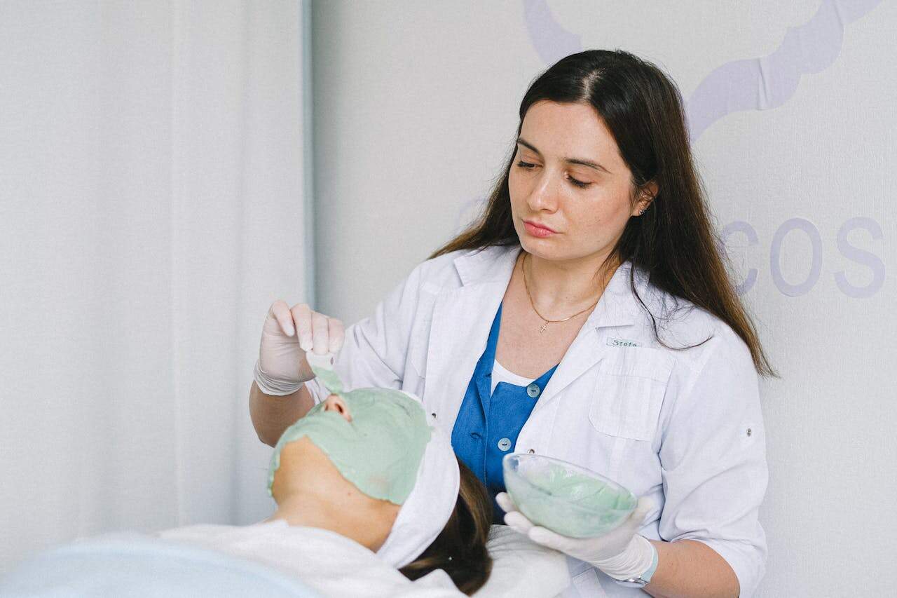 A beautician applies a green facial mask to a client in a wellness clinic, promoting skin care and relaxation.