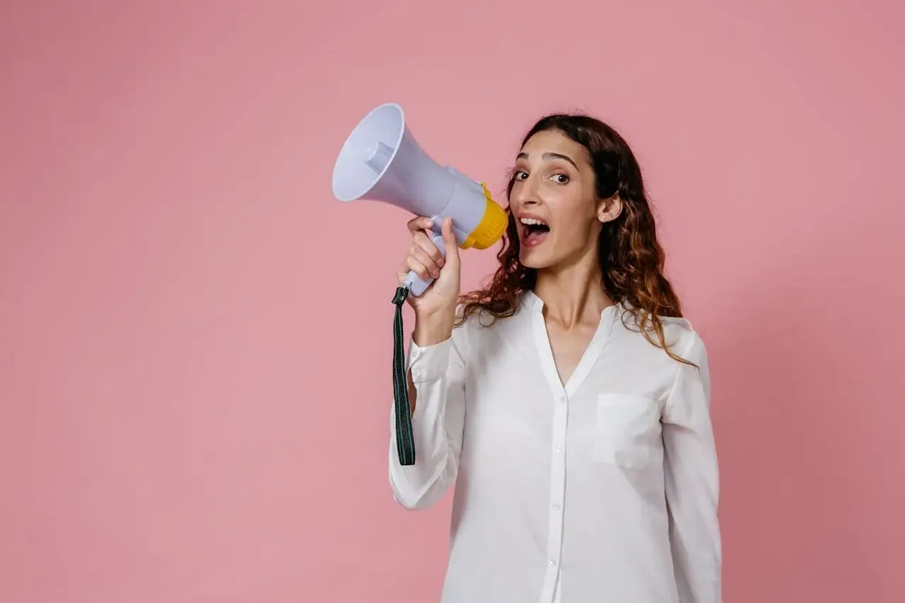 Mastering the First Impression: Your intriguing post title goes here Woman in white shirt holding megaphone against a pink background, confidently speaking.